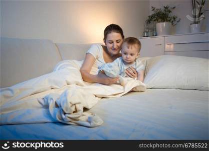 Cute baby boy and his mother playing with toy on bed before going to sleep