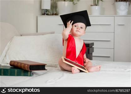 Cute baby boy in academician mortarboard sitting on bed and looking in camera