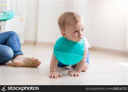Cute baby boy in apron crawling on floor while mother trying to feed him