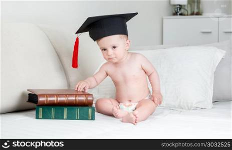 Cute baby boy in diapers and graduation cap sitting on bed with books. Baby boy in diapers and graduation cap sitting on bed with books
