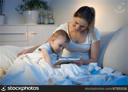 Cute baby boy sitting on bed with mother and using digital tablet at night