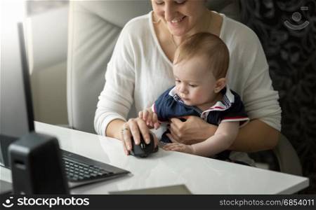 Cute baby boy sitting on mothers lap and playing with computer mouse