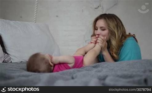 Cute baby girl lying on bed while her caring mother expressing her love, tenderness and care, kissing newborn&acute;s feet. Affectionate mom nursing infant child. Beautiful woman holding and kissing baby&acute;s feet. Slow motion. Steadicam stabilized shot.