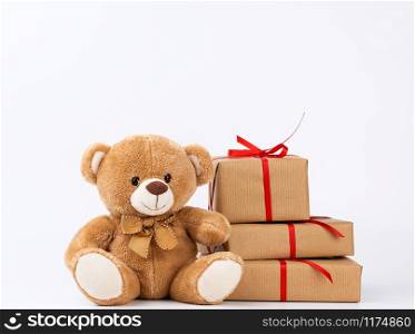 cute beige teddy bear and stack of gifts in boxes wrapped in brown eco paper, tied with silk red ribbon on a white background, happy birthday and valentines day