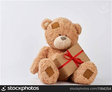 cute big beige teddy bear holds a brown box with a red ribbon on a white background. Festive concept