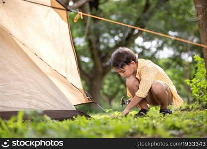 Cute boy holding hammer setting up a tent on grassy field in forest at weekend.