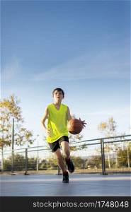 Cute boy in yellow shirt plays basketball on city playground. Active teen enjoying outdoor game with orange ball.