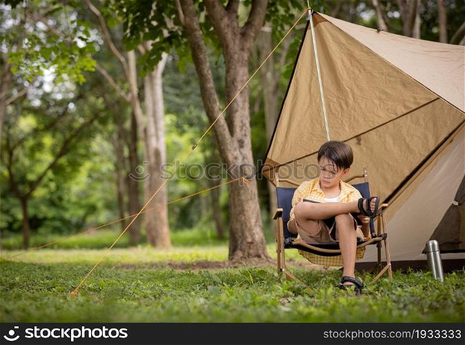 Cute boy sitting intently reading a book in front of a tent during camping.