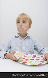 Cute boy with cake slice in plate on table looking away at home