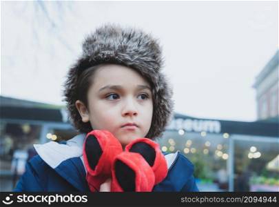 Cute boy with fluffy hat and warm jacket walking outside in the city, Kid with thinking face standing outside with blurry street background,Child having fun playing in old town on Winter holiday