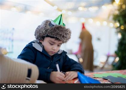 Cute boy with party hat using pink pen drawing or writing on paper colour with blurry bright light background, Kid having fun at birthday party, Child doing activity on Christmas or New Year holiday