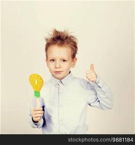 Cute boy with yellow paper lightbulb against a white background. little student gesturing thumb holding toy bulb. Cheerful smiling Kid with funny photo props. School concept. Back to School