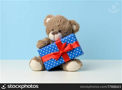cute brown teddy bear holding a box wrapped in blue paper and red silk ribbon on white table. Prize and congratulations