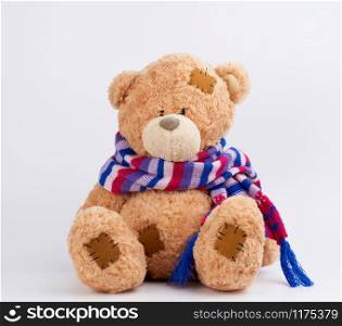 cute brown teddy bear with patches in a colored knitted scarf sitting on a white background, close up
