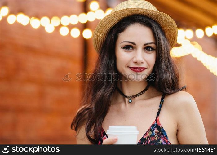 Cute brunette female with dark eyes, red lips and pure healthy skin, wearing straw hat and dress holding takeaway coffee posing at terrace. Young female having coffee break after hard work at office
