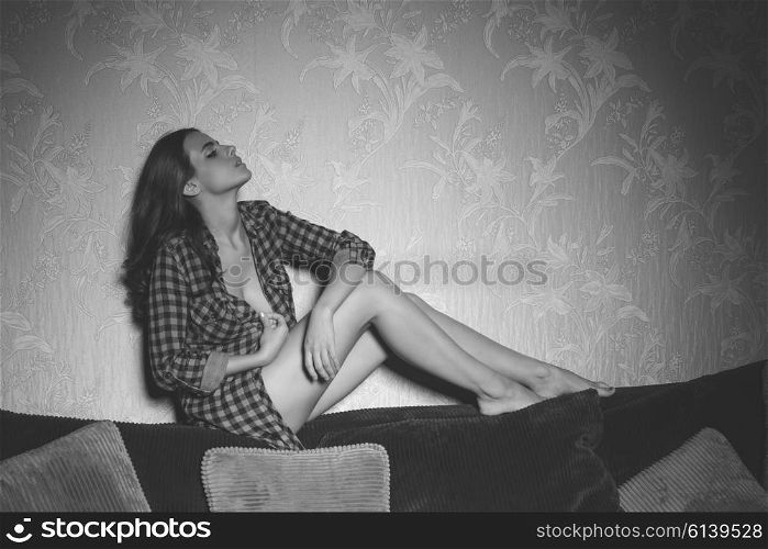 cute brunette woman with long hair in sexy pose on backrest of sofa with nude legs and unbottoned plaid shirt, black and white image