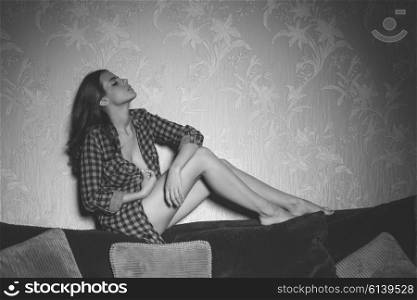 cute brunette woman with long hair in sexy pose on backrest of sofa with nude legs and unbottoned plaid shirt, black and white image