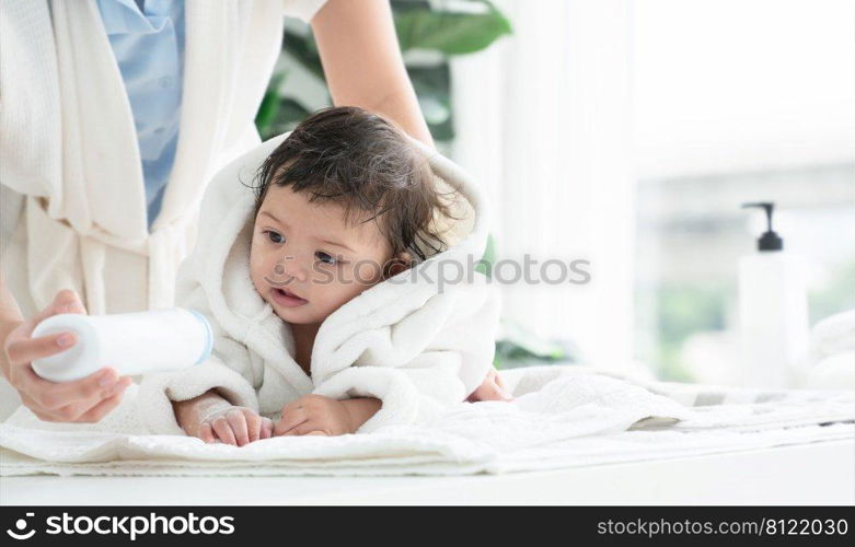 Cute Caucasian litt≤todd≤r baby girl wear bathrobe after bathing is smiling and lying on towel whi≤mother wipe and apply talcum powder on her body at home. Hygie≠care forχldren concept