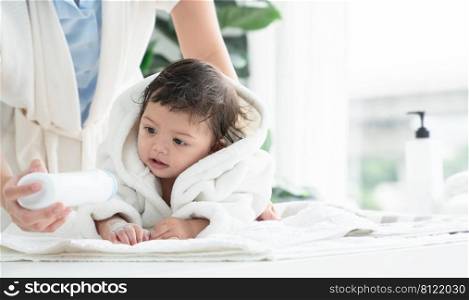 Cute Caucasian litt≤todd≤r baby girl wear bathrobe after bathing is smiling and lying on towel whi≤mother wipe and apply talcum powder on her body at home. Hygie≠care forχldren concept