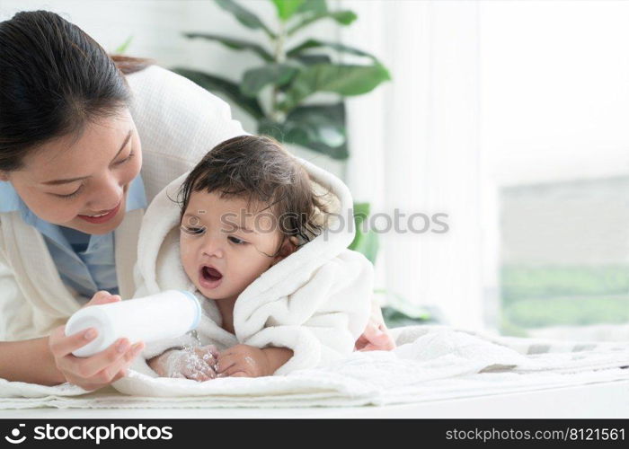 Cute Caucasian little toddler baby girl wear bathrobe after bathing is smiling and lying on towel while mother wipe and apply talcum powder on her body at home. Hygiene care for children concept