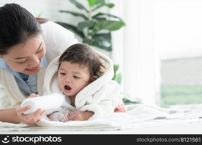 Cute Caucasian little toddler baby girl wear bathrobe after bathing is smiling and lying on towel while mother wipe and apply talcum powder on her body at home. Hygiene care for children concept