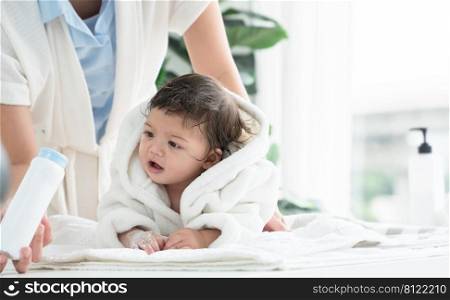 Cute Caucasian little toddler baby girl wear bathrobe after bathing is smiling and lying on towel while mother wipe and apply talcum powder on her body at home. Hygiene care for children concept