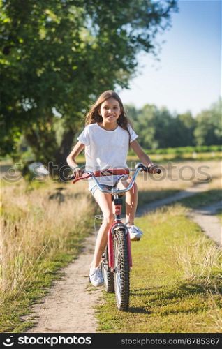 Cute cheerful girl with long hair riding her bicycle on dirt road at meadow