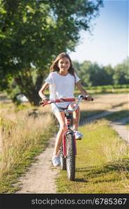 Cute cheerful girl with long hair riding her bicycle on dirt road at meadow