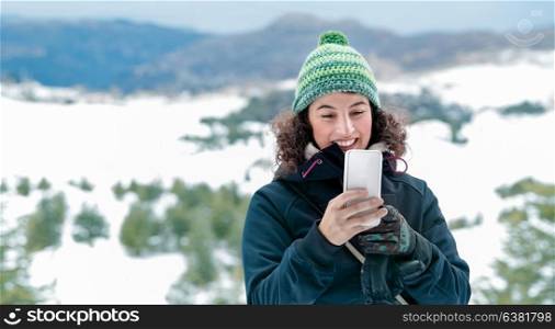 Cute cheerful woman using mobile phone in the mountains, receiving funny messages, having fun outdoors, with pleasure spending winter vacation in the snowy mountains