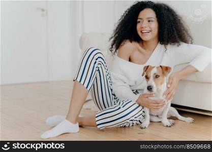Cute cheerful young African American female sits on floor near sofa, plays with dog, dressed in stylish clothing, looks gladfully somewhere, spends free time at home. Happy pet owner indoor.