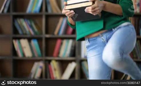 Cute college female student carrying a pile of books in library. Mid section of hipster girl looking through many books while sitting on wooden bench in library with books for self-education. University student studying hard to prepare for exams.