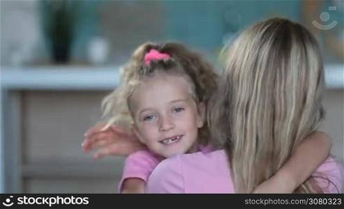 Cute curly blonde daughter with ponytail embracing her loving mother in domestic interior and smiling. Adorable little girl expressing her love and tenderness to beautiful mom with hugs and kisses.