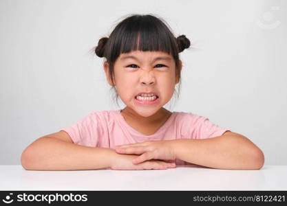 Cute dark haired little girl with angry face sitting at a table on a white background and looking at the camera.