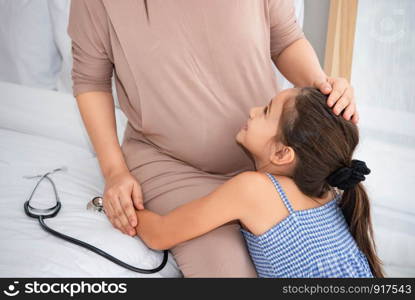 Cute daughter hugging mother and listening to voice of baby in mother belly for checking and looking at mom face. Pregnant and parents. Health and medical. People lifestyles and family love concept.