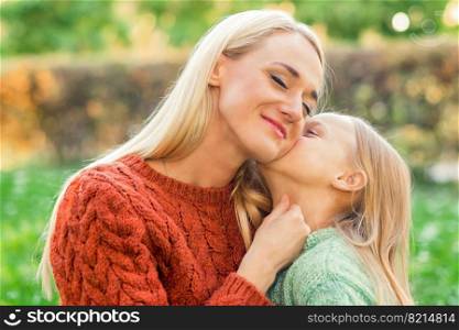 Cute daughter kisses her young mother while sitting at the park. Daughter kisses her young mother