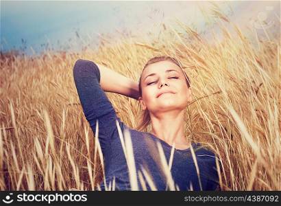 Cute dreamy girl on golden ripe wheat field, lying down with closed eyes and enjoying beautiful mild sunset light and autumn nature