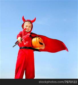 Cute Emotional Little Boy in a Costume of Red Devil Standing with a Trident and Jack O&rsquo; Lantern at the Blue Sky.