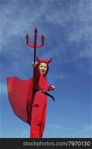 Cute Emotional Little Boy in a Costume of Red Devil Standing with a Trident at the Blue Sky