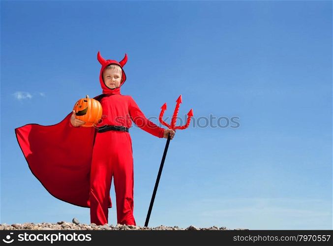 Cute Emotional Little Boy in a Costume of Red Devil Standing with a Trident and Jack O&rsquo; Lantern at the Blue Sky.
