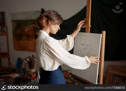 Cute female artist drawing in studio. Creative paint, pencil sketch on easel, workshop interior on background