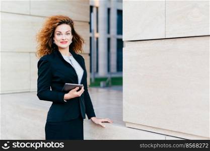 Cute female freelancer with luxurious light hair, blue eyes, red rouged lips and pure skin, wearing black formal suit, holding electronic gadget n hands, going on meeting with her business partners
