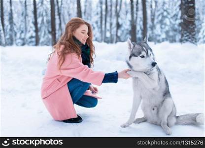 Cute female master on dog training with siberian husky, snowy forest on background. Cute girl with funs with charming pet. Female master on dog training with siberian husky