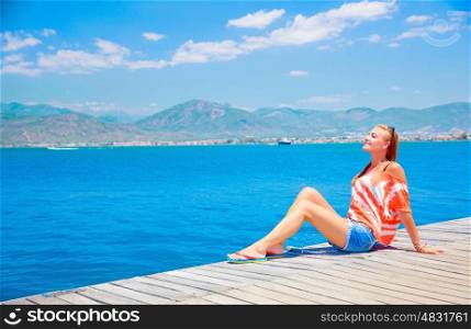 Cute female sitting on wooden pier and enjoying bright sun light, spending warm sunny day on the beach, summertime leisure concept