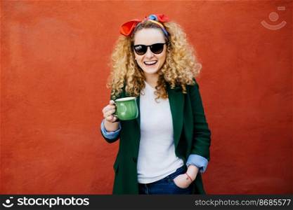 Cute female student with curly blonde hair wearing headband, sunglasses and stylish jacket holding cup of coffee looking at camera with sincere smile. Beautiful woman enjoying drinking tasty tea