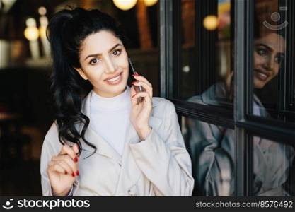 Cute female with wavy dark hair, charming eyes, well-shaped lips and white even teeth wearing white clothes having red manicure communicating over telephone touching her hair with her gentle hand