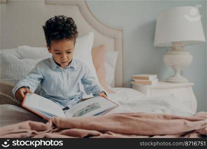 Cute focused small boy in casual wear holds and tries to read childrens book, sitting on bed with nightstand next to with white elegant l&on it. Kid spending weekend morning leisure at home. Cute afro american small boy trying to read children’s book