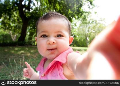 Cute funny happy baby infant sticking arm out as if she?s taking self portrait photo in a park.