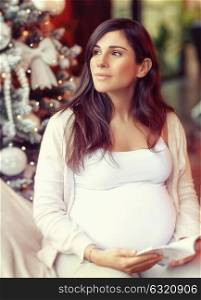 Cute gentle pregnant woman sitting near beautiful decorated Christmas tree and reading book, warm Christmastime at home