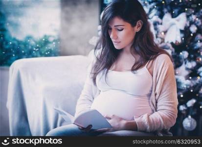Cute gentle pregnant woman sitting near beautiful decorated Christmas tree at home and reading book, telling interesting fairy tale for her future baby