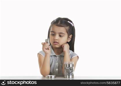 Cute girl holding Indian coin while sitting at table against white background